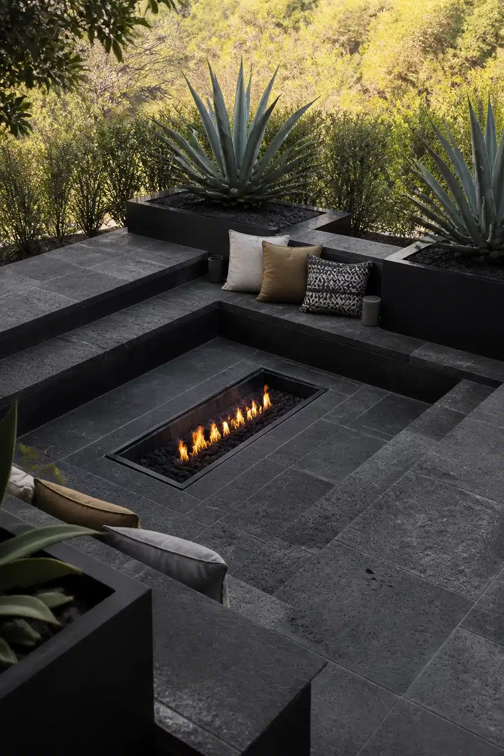 Modern sunken fire pit with dark basalt stone pavers, built-in seating, and architectural agave plants outdoors.