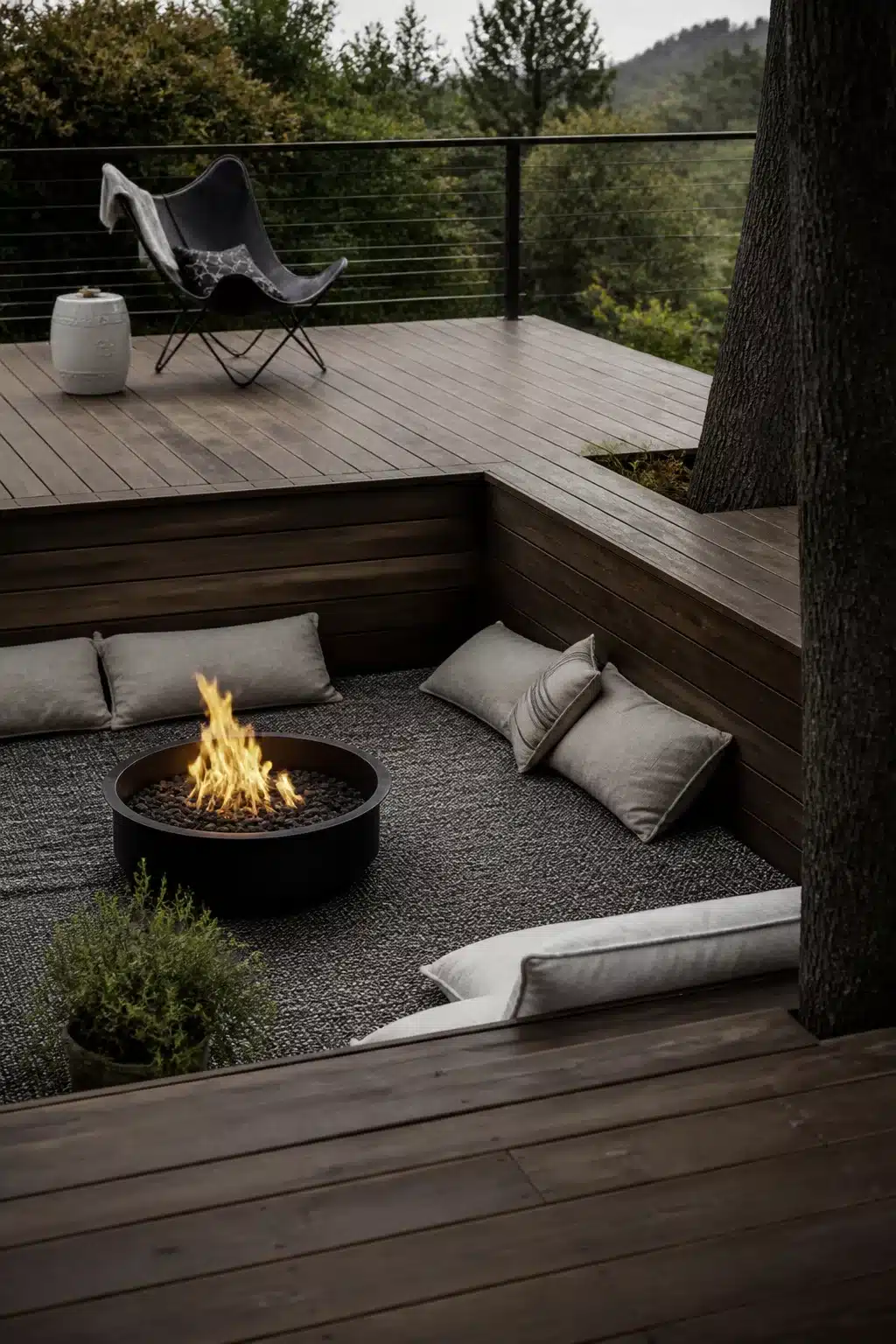 Sunken fire pit lounge with round black fire bowl on a split-level dark timber deck surrounded by grey cushions.
