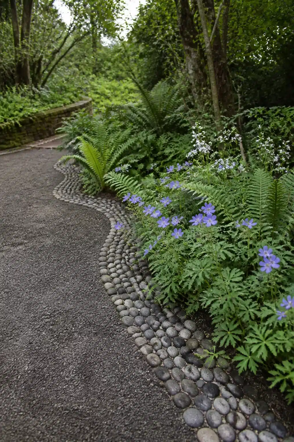 River pebble mosaic edging alongside ferns and blue geraniums — woodland garden edging ideas with natural stone.