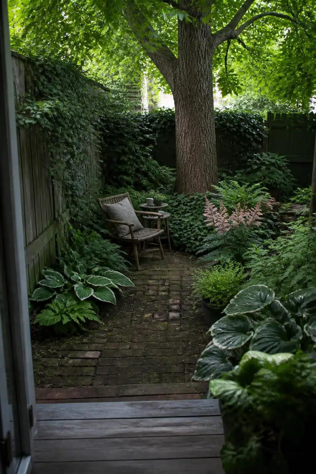 Narrow city woodland shade garden with brick path, rattan chair, hostas and ferns under a dense tree canopy.