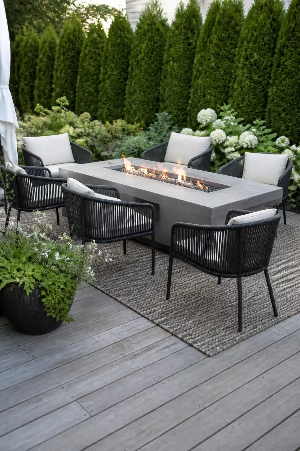 Concrete fire pit dining table with rope chairs on a gray deck — backyard firepit landscaping ideas with arborvitae hedge