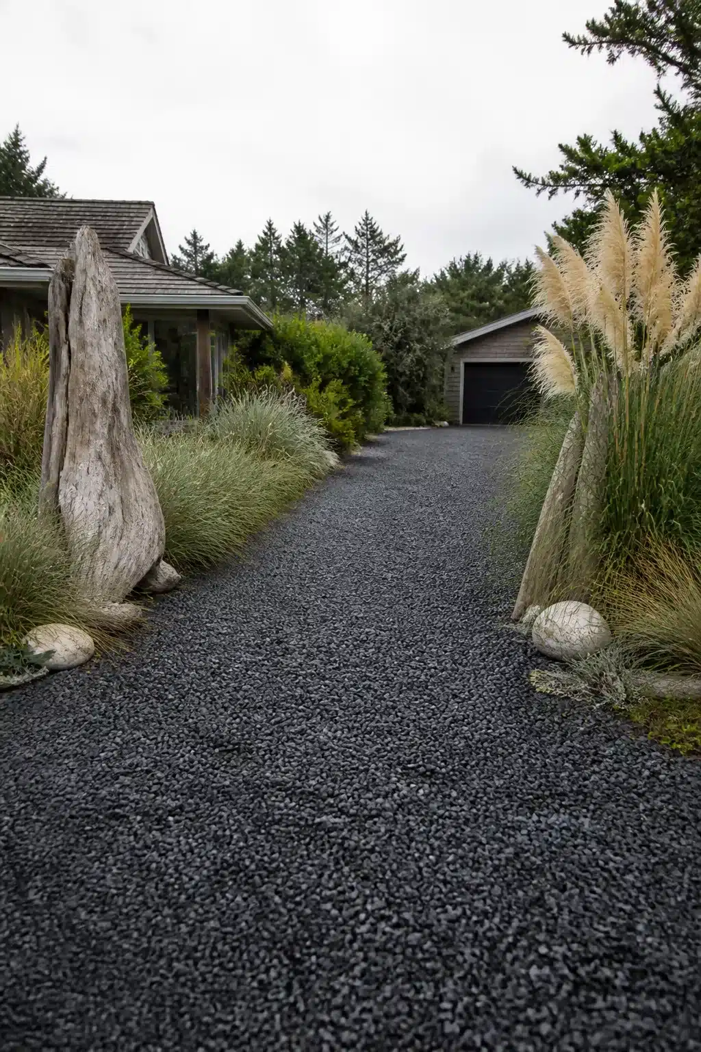 Dark black gravel driveway ideas with driftwood sculptures and pampas grass in a coastal garden setting