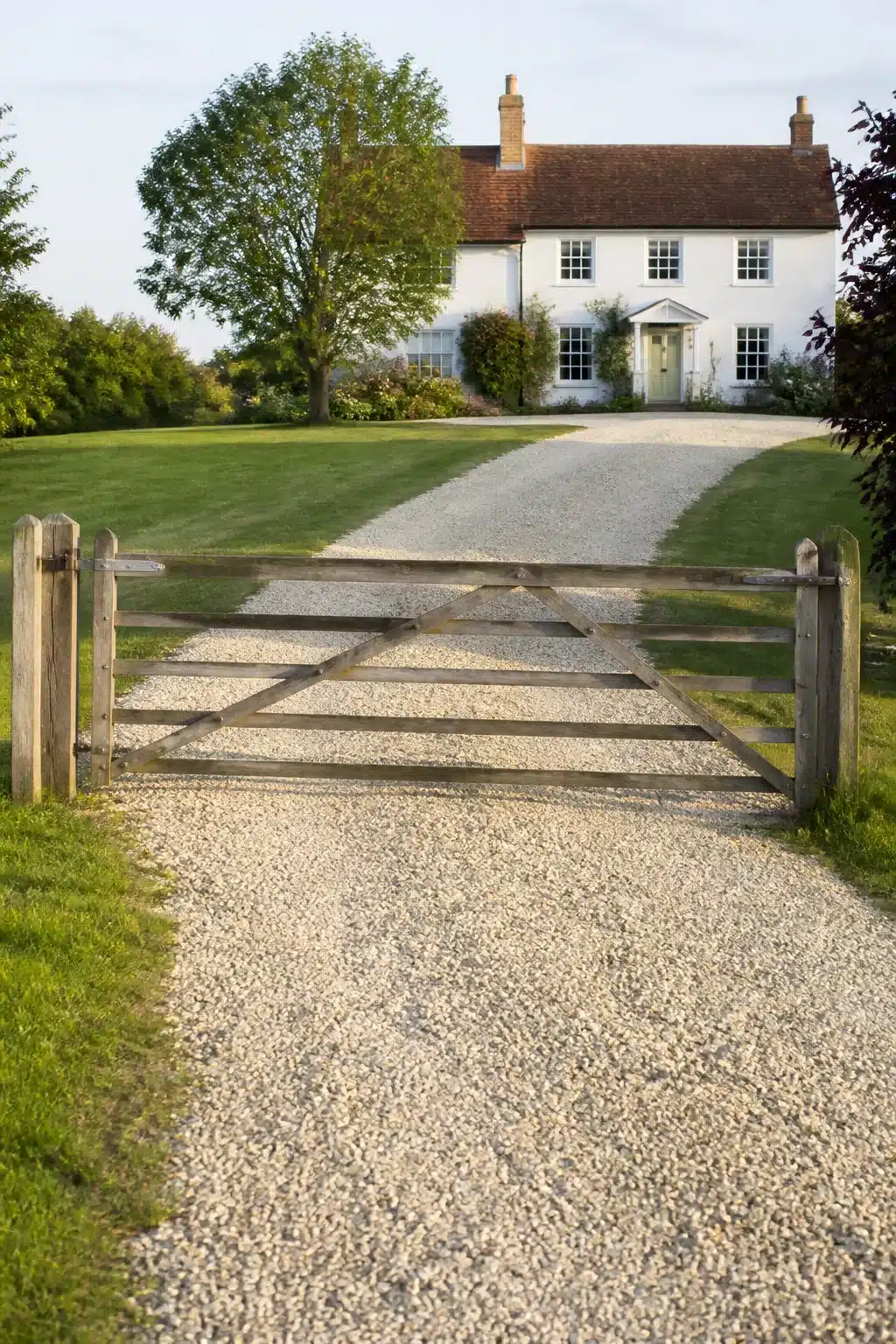 Weathered timber five-bar gate opening onto a pale crushed gravel driveway at a white farmhouse — Gravel Driveway Ideas
