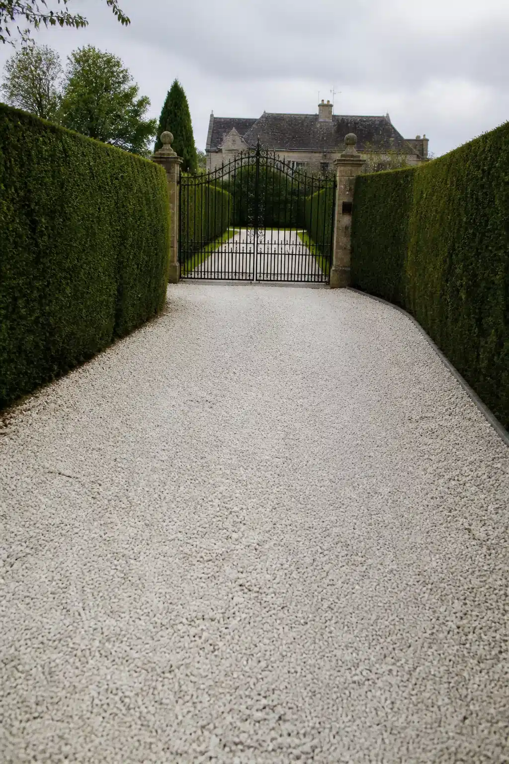 White quartz gravel driveway ideas with tall yew hedges and wrought iron estate gate leading to stone manor