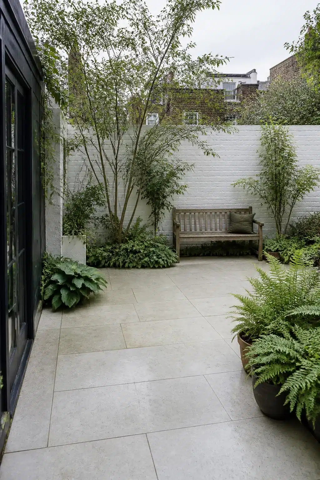 North facing garden ideas with pale limestone-look porcelain paving, shade planting, and a timber bench in a courtyard garden.