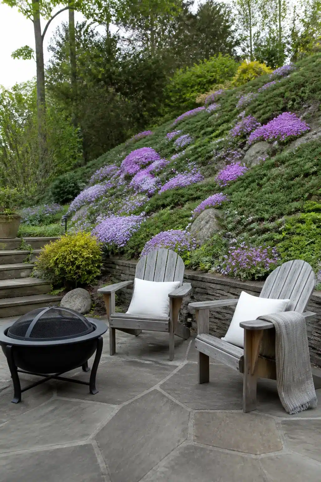 Sloped backyard landscaping ideas with creeping phlox hillside, flagstone patio, and Adirondack chairs by fire pit