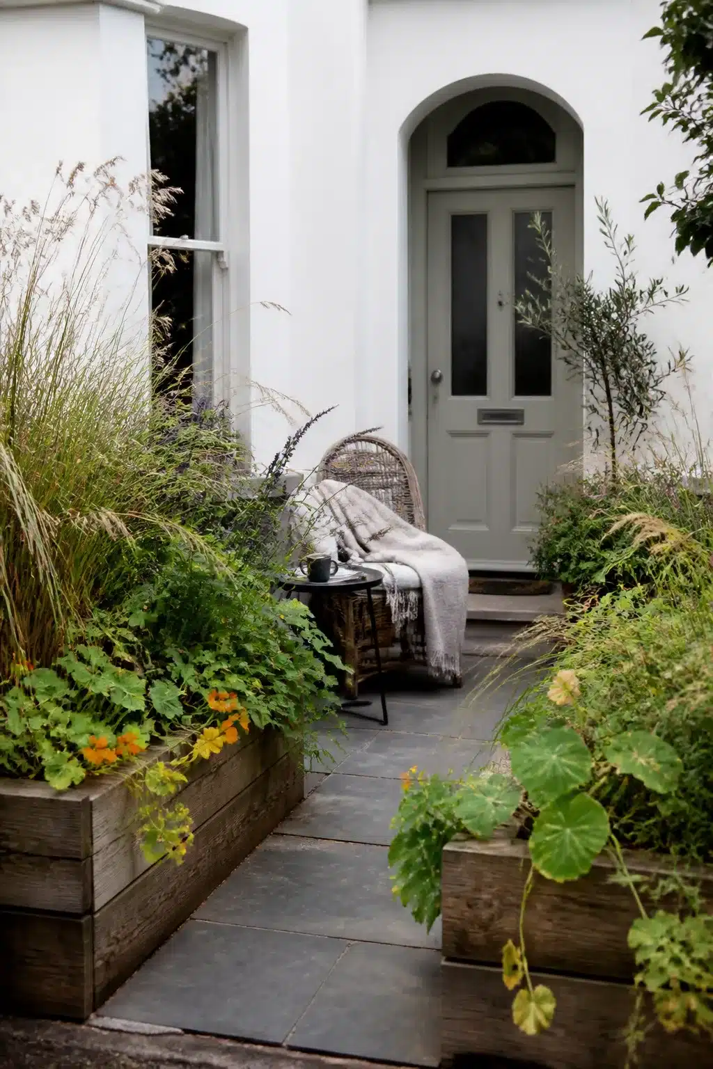 Small front garden with weathered timber raised beds, slate paving path, and arched sage-green door