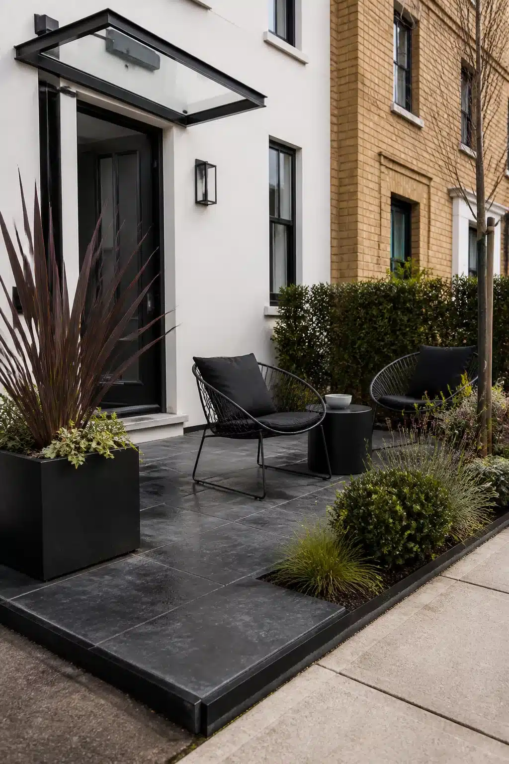 Small front garden with dark slate paving, black wire chairs, and phormium planting outside a modern white house entrance.
