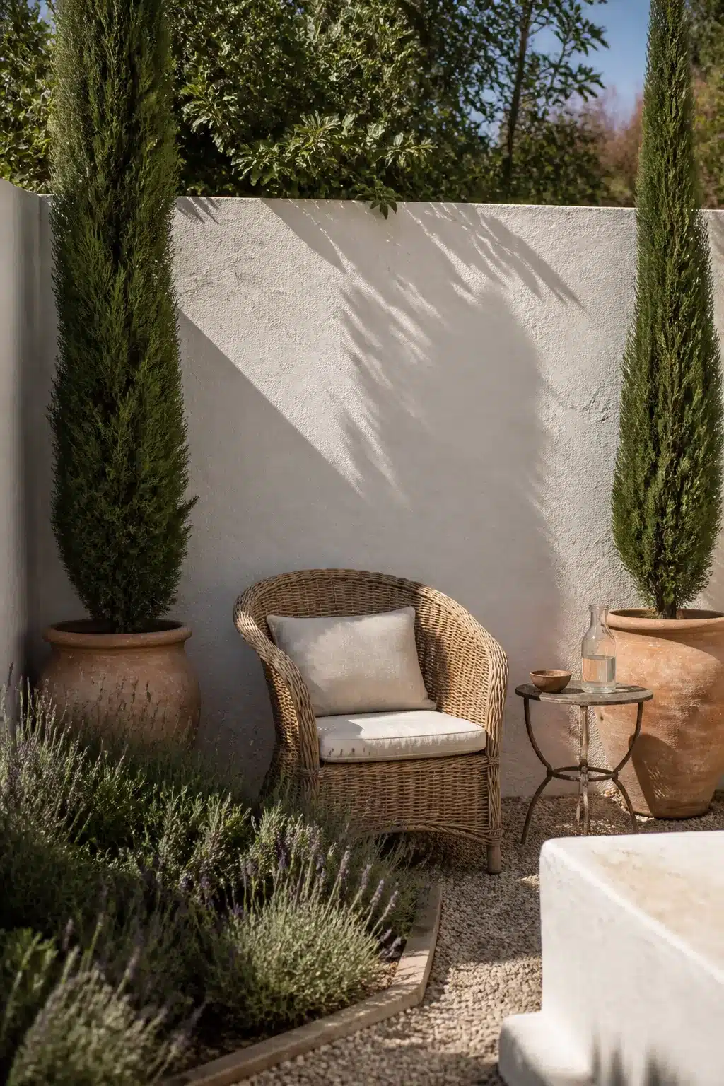 Slender Italian cypress trees in terracotta urns flank a wicker chair in a small Mediterranean garden courtyard.
