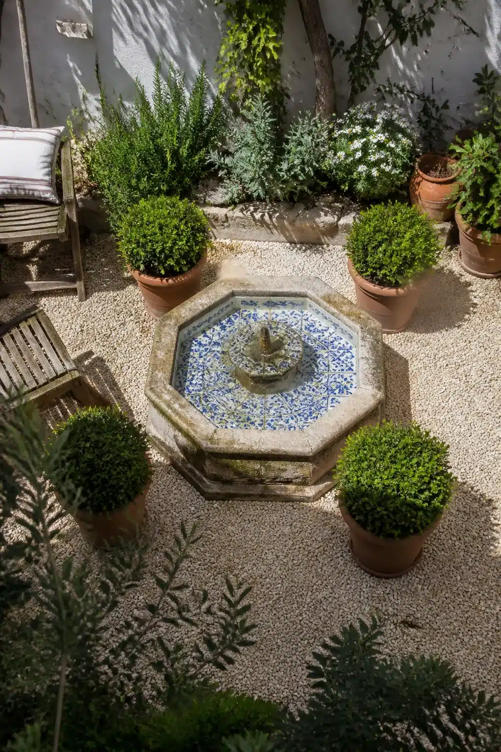 Octagonal stone fountain with blue mosaic tiles surrounded by clipped boxwood in terracotta pots — small Mediterranean garden courtyard