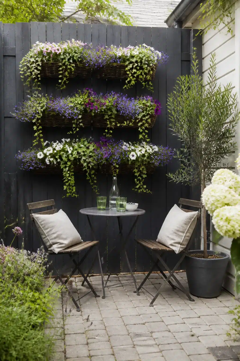 Small patio garden with stacked wicker wall baskets of trailing flowers on a dark fence and bistro seating