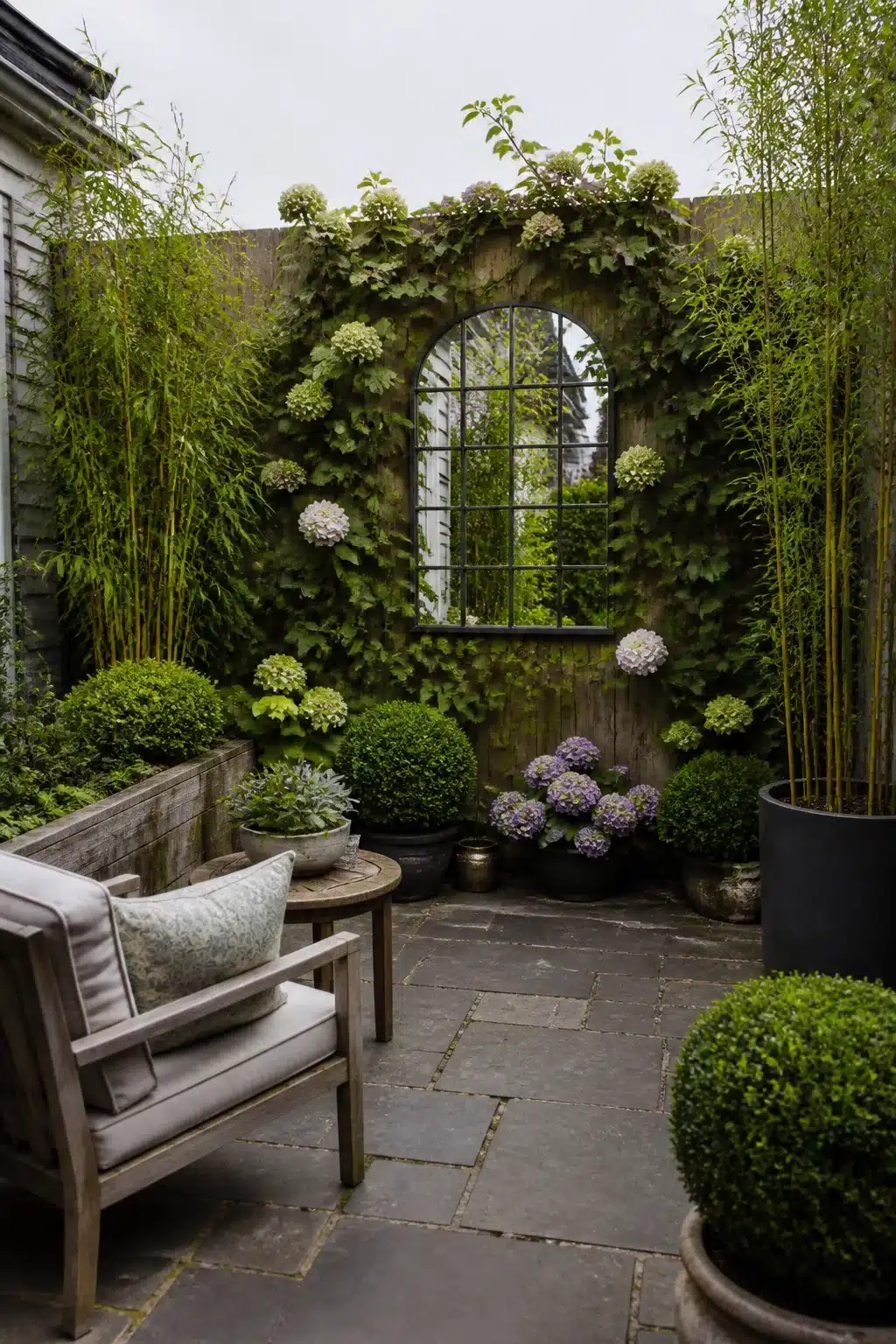 Small patio garden with arched mirror on stone wall, hydrangeas, box balls, and bamboo in containers