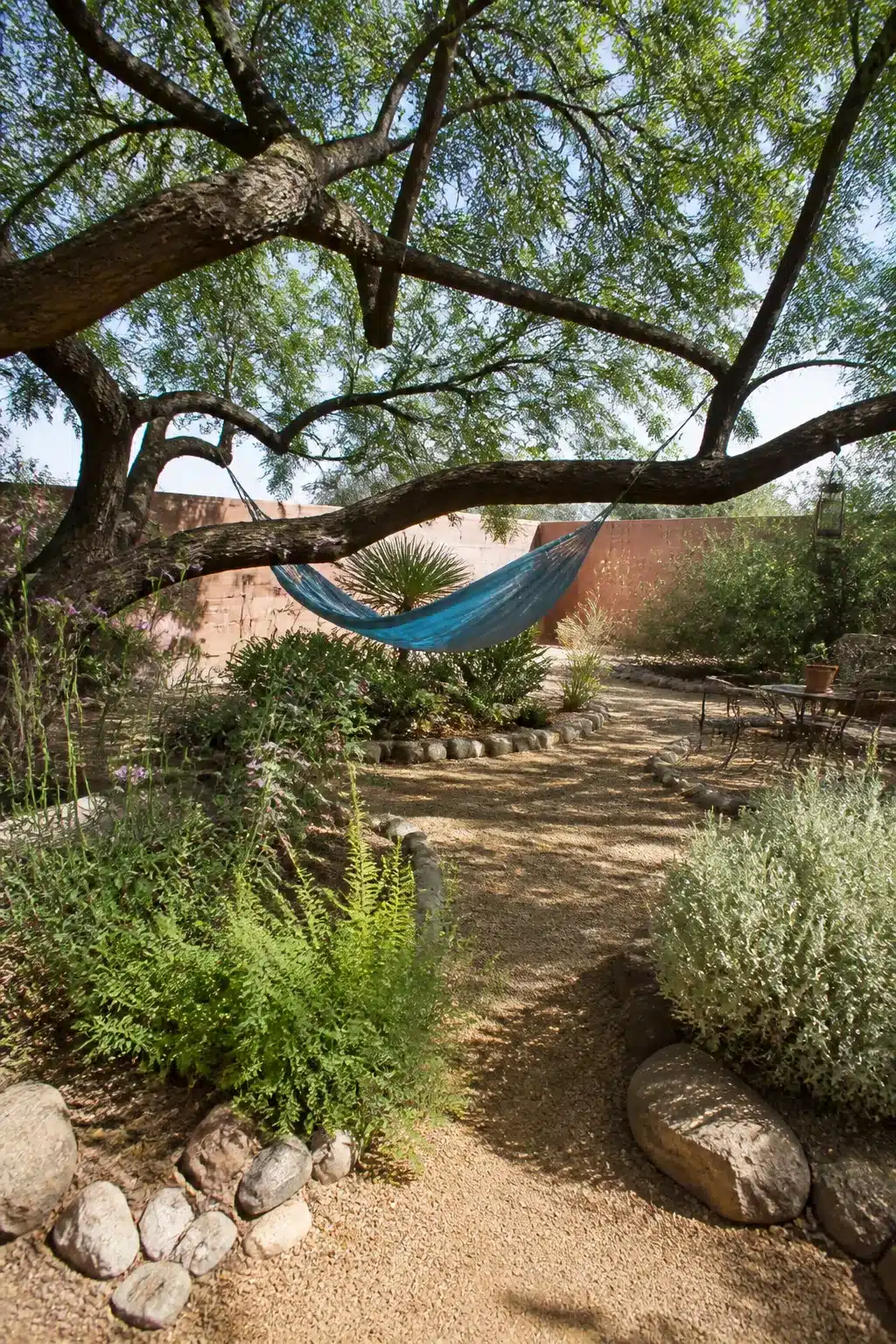 Xeriscape backyard with hammock under mesquite tree, gravel path, and drought-tolerant plants edged with river rocks.