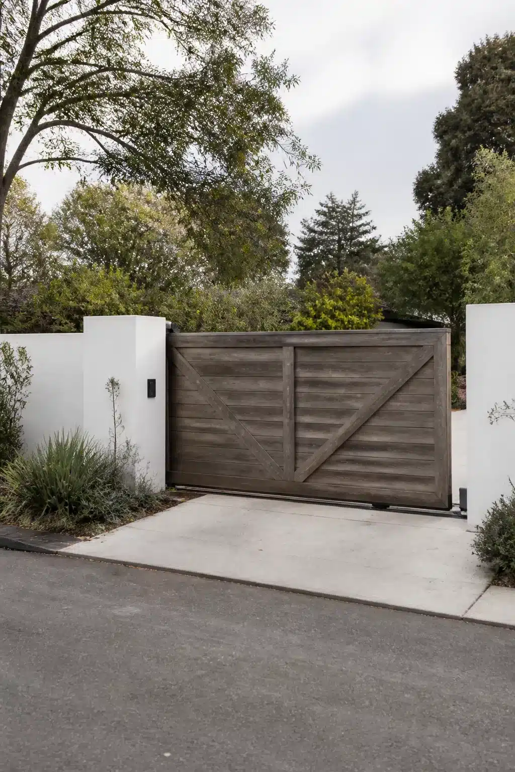 Gray-stained sliding barn-style wooden driveway gate ideas with white rendered pillars and concrete entrance
