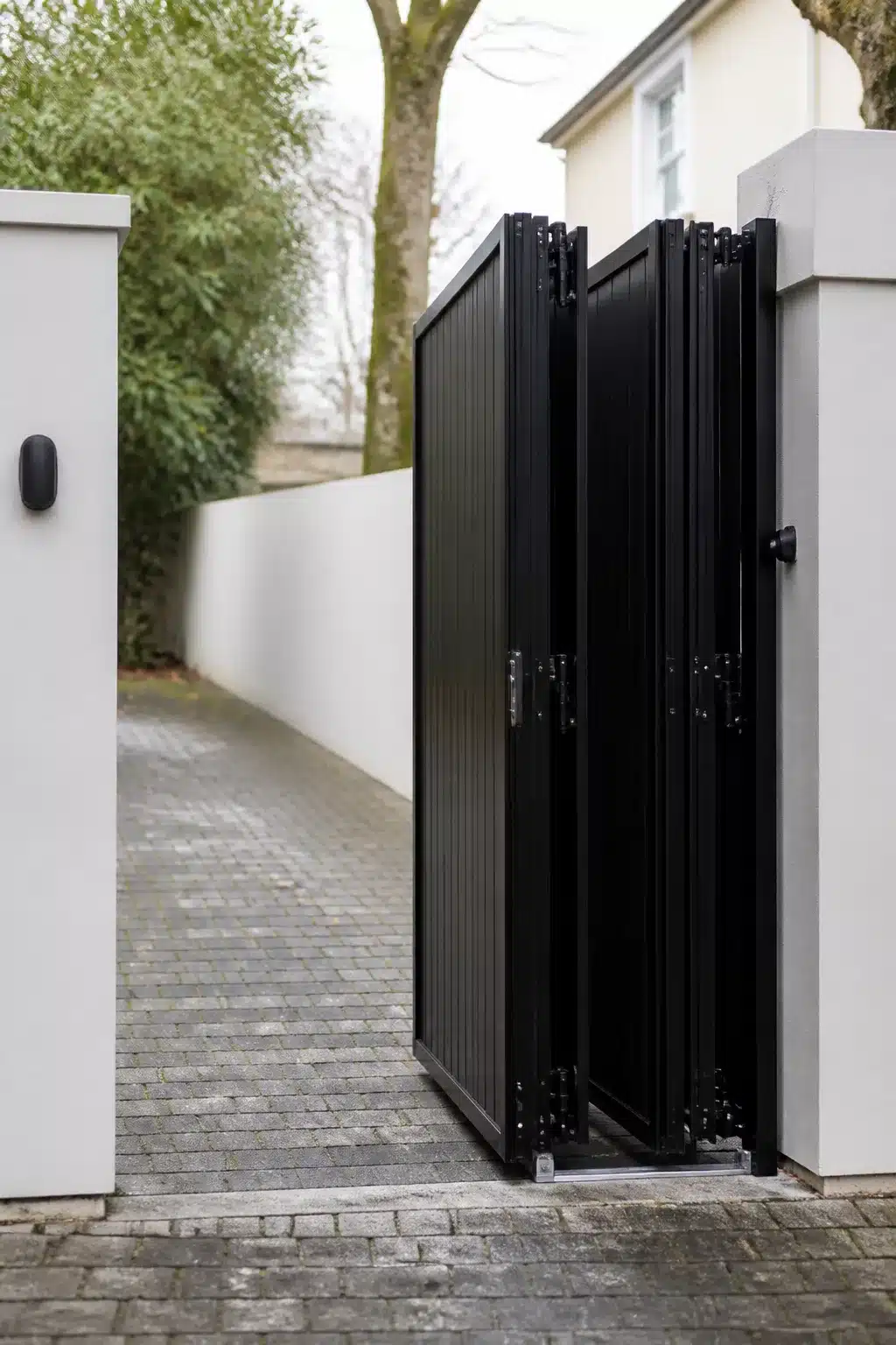 Automated black aluminium bi-fold driveway gate ideas with white rendered pillars and cobblestone entrance