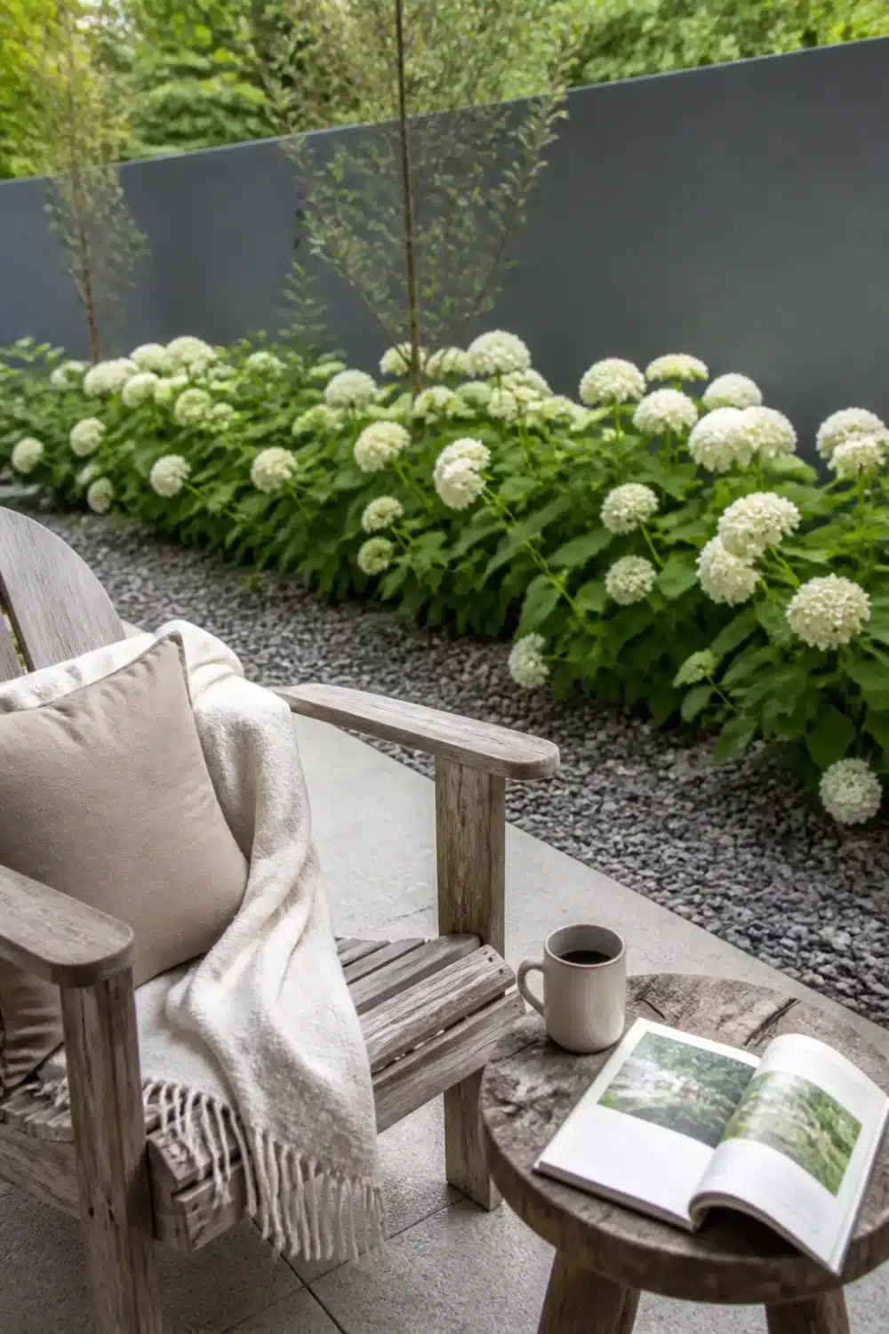 White hydrangea landscaping strip against a dark gray garden wall with gravel mulch and a teak chair